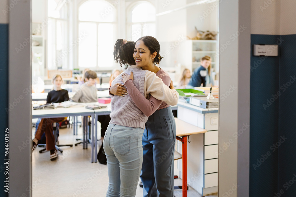 Schoolgirl embracing female teacher standing in classroom Schoolgirl embracing female teacher standing in classroom