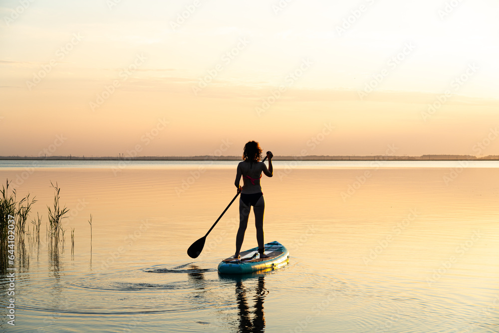 Sports girl on board for glanders surfing. young slender woman floats on a board with a paddle. Surfing. Summer fun on the water. Healthy lifestyle. Woman stand up paddle boarding SUP on calm water - obrazy, fototapety, plakaty 