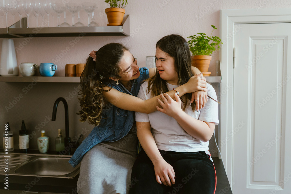 Smiling young woman hugging sister with down syndrome in kitchen at home Smiling young woman hugging sister with down syndrome in kitchen at home