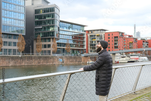 young bearded man in coat and hat stands on shore river Main, winter walks in Frankfurt, View of modern residential buildings, Gutleitviertel district, boats and yachts moored on banks photo