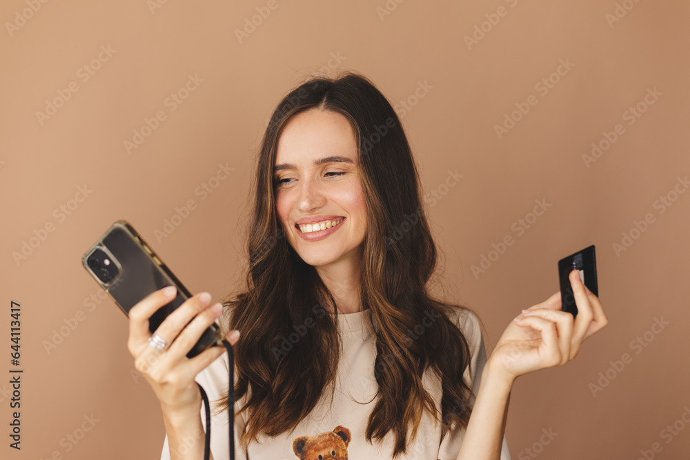 Online shopping. Portrait of smiling brunette curly hair woman paying with plastic credit card on smartphone app, standing with mobile phone and bank card against beige background. Happy emotions. Online shopping. Portrait of smiling brunette curly hair woman paying with plastic credit card on smartphone app, standing with mobile phone and bank card against beige background. Happy emotions.