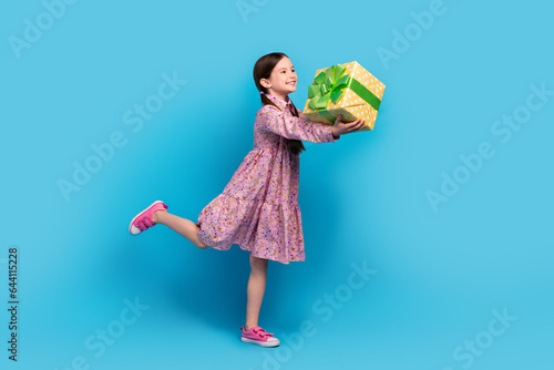 Full body length photo of funny small girl kid send you wrapped yellow package surprise celebrate birthday isolated on blue color background