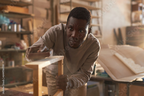 Front view portrait of young black man sanding wooden furniture in carpentry workshop, copy space