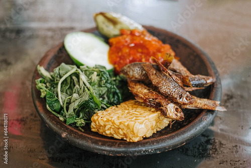 A wooden plate of Indonesian traditional food called Lalapan contains fried Iwak Wader or Spotted Barb fish, vegetables, fried tempe, cucumber and egg plant with sambal or chili. Selective focus. photo