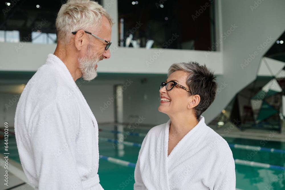 Obraz premium joyful middle aged couple in glasses and white robes chatting near indoor pool in spa center