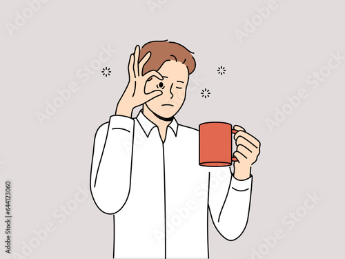 Sleepy man with mug of coffee, opens eyes with hand and needs to rest due to lack of energy and drowsiness. Tired young guy drinks coffee drink to cheer up and continue productive work.