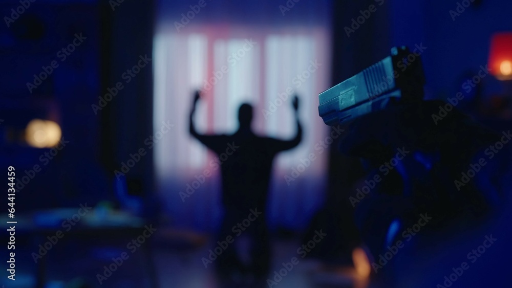 The arrest of a burglar in a dark apartment lit by blue light. The offender is on his knees, with his hands up. A gun in the hand of a policeman on a blurred background of a criminal close up. The arrest of a burglar in a dark apartment lit by blue light. The offender is on his knees, with his hands up. A gun in the hand of a policeman on a blurred background of a criminal close up.