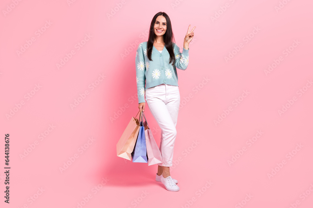 Full length photo of shiny sweet woman dressed blue cardigan showing v-sign holding shoppers isolated pink color background Full length photo of shiny sweet woman dressed blue cardigan showing v-sign holding shoppers isolated pink color background