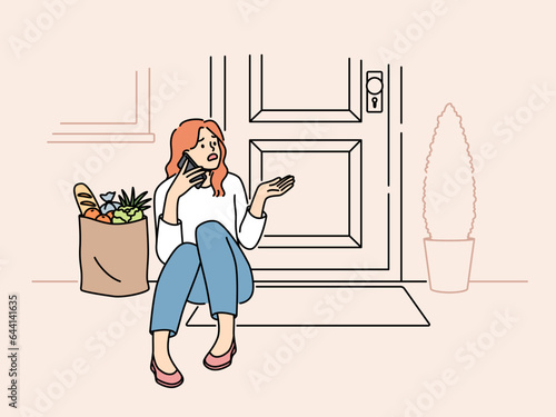 Embarrassed woman sits on threshold near door with jammed lock and cannot get into house, makes phone call. Puzzled girl with bag of groceries, calling specialist because of loss of key to door