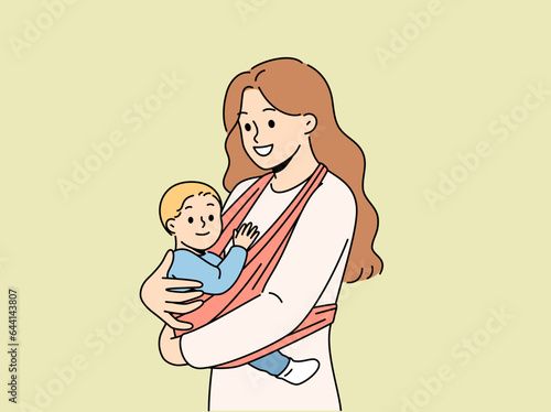 Mother holds baby in carrier sling and smiles, taking care of son and using comfortable babywearing. Caring happy woman with newborn, recommends purchasing sling for young mothers.
