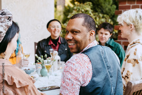 Portrait of smiling gay man looking over shoulder by friends during dinner party in back yard photo