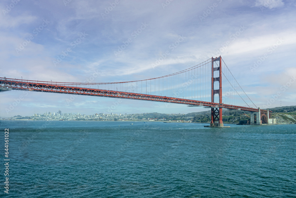 Fototapeta premium San Francisco, CA, USA - July 13, 2023: Golden Gate bridge south landing , ocean side, with 1 tower under blue cloudscape. Green hills and city on horizon under the span