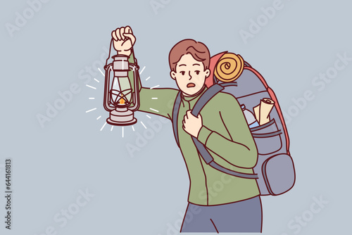 Man is engaged in hiking and carries large backpack on back, holding kerosene lantern to light way in dark. Frightened guy hears rustle in bushes and looks around while hiking in forest