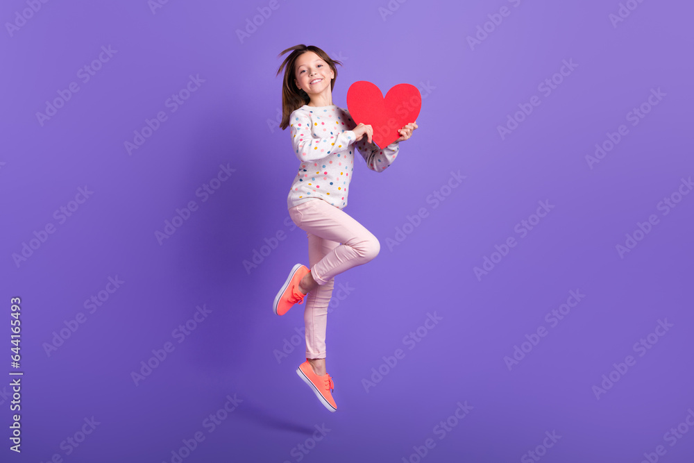Full length photo of pretty shiny little girl dressed dotted pullover jumping high holding red heart isolated purple color background Full length photo of pretty shiny little girl dressed dotted pullover jumping high holding red heart isolated purple color background