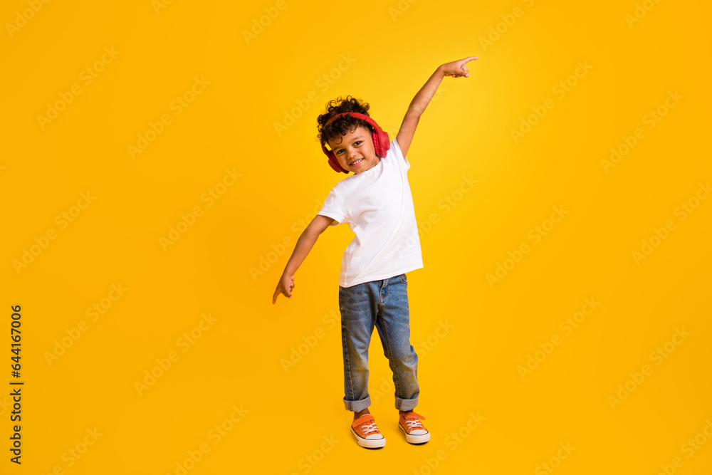 Full length photo of cheerful cool little boy dressed white t-shirt pointing fingers enjoying songs earphones isolated yellow color background Full length photo of cheerful cool little boy dressed white t-shirt pointing fingers enjoying songs earphones isolated yellow color background