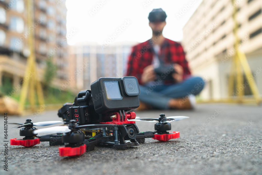 Close up shot of generic desing fpv drone after landing on a street road, with male pilot  making aerial urban photography and videography on the background