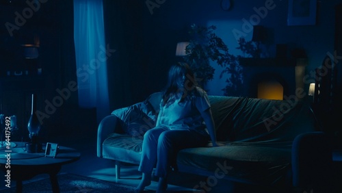 Shot capturing a young woman sitting on the couch in a dark room. Ominious, spooky, creepy atmosphere. Something paranormal is about to happen. photo