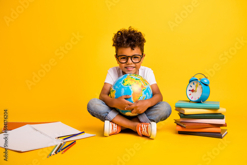 Full body photo of clever positive small kid sit floor hold world globe pile stack book clock notebook isolated on yellow color background