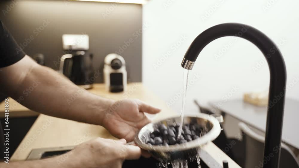 A man washes fresh blueberries under running water in a colander. Close-up. A man washes fresh blueberries under running water in a colander. Close-up.