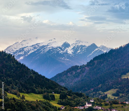 Summer Alps mountain landscape with village, fir forest on slope and snow covered rocky tops in far, Austria.