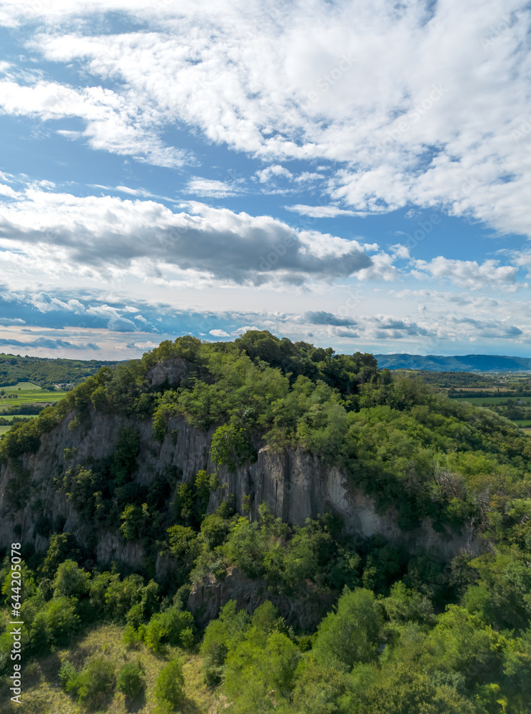 Obraz premium Aerial view of steep cliff on a hill with green trees growing on top below blue sky with clouds in Italian countryside landscape