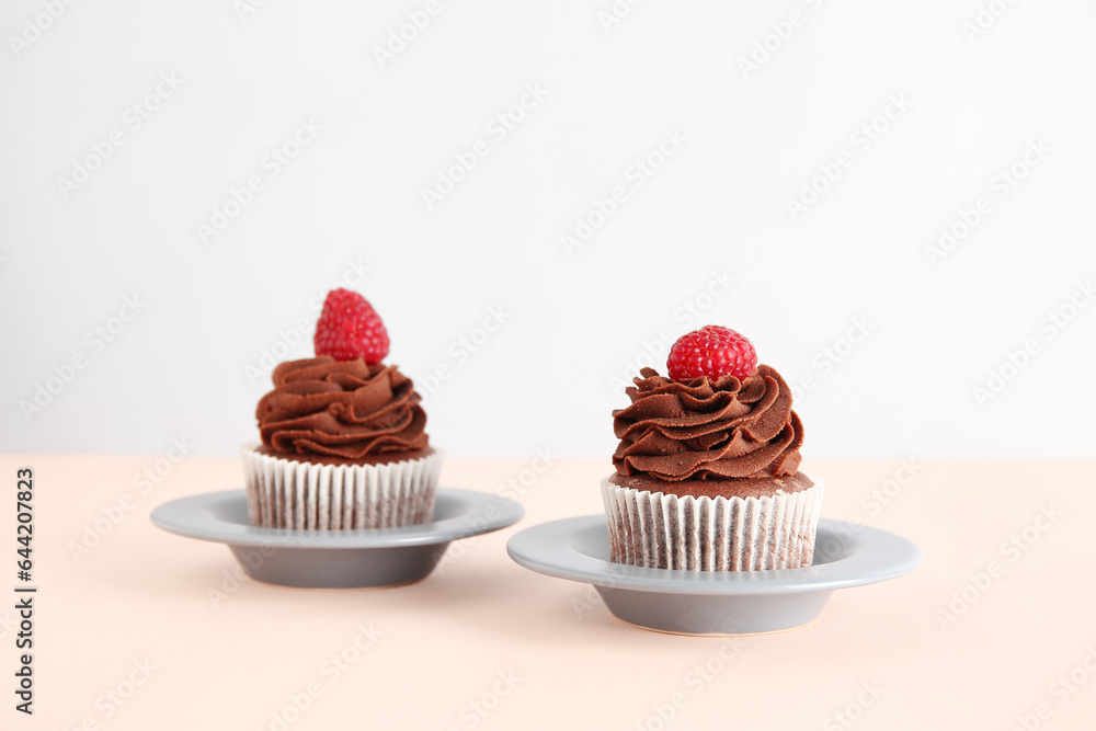 Plates of tasty chocolate cupcakes with raspberries on beige background Plates of tasty chocolate cupcakes with raspberries on beige background