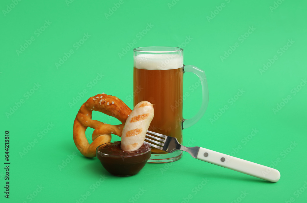 Fork with tasty Bavarian sausage, pretzel, sauce and mug of beer on green background. Oktoberfest celebration Fork with tasty Bavarian sausage, pretzel, sauce and mug of beer on green background. Oktoberfest celebration