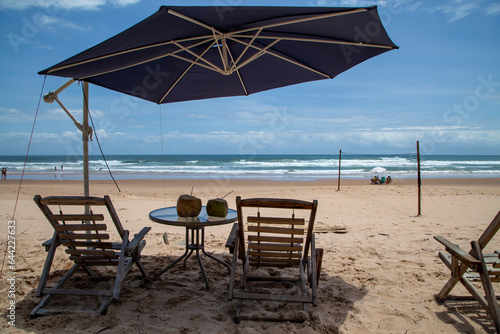 Cocos com canudinho na mesa e cadeiras de praia vazias esperando turistas em praia ensolarada do nordeste brasileiro  com mar e ondas no horizonte