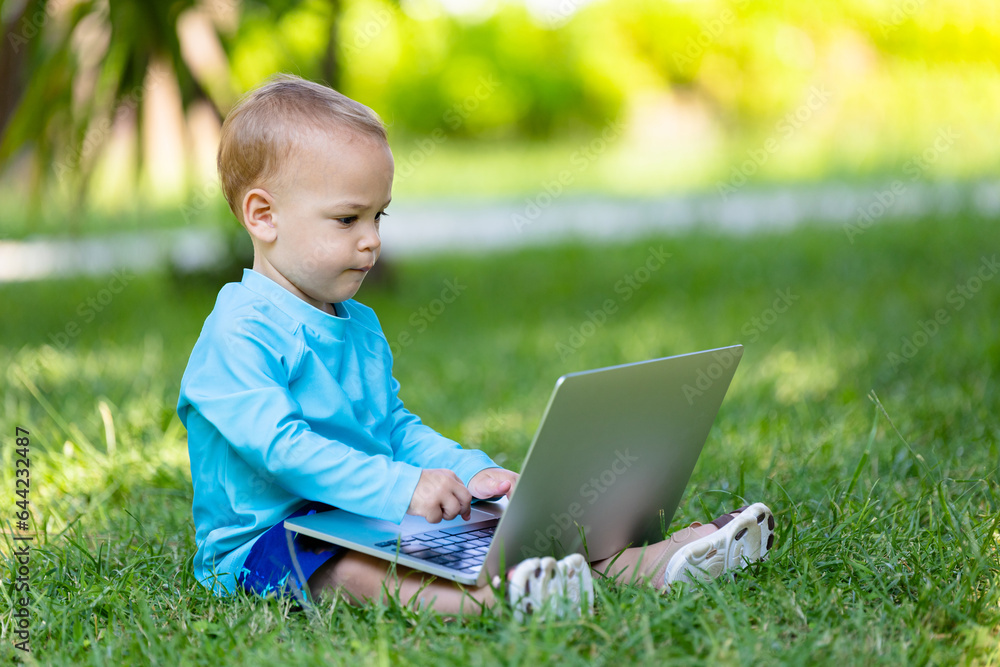 Little baby boy with hands on computer keyboard looking at laptop screen in nature. Kids and technology vacation concept.