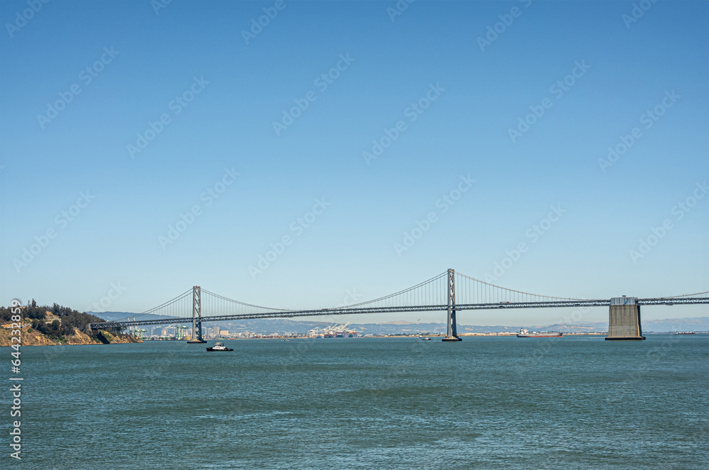 Fototapeta premium San Francisco, CA, USA - July 13, 2023: SF-Oakland Bay Bridge lands on Yerba Buena Island under blue sky above blue water. Ships on horizon