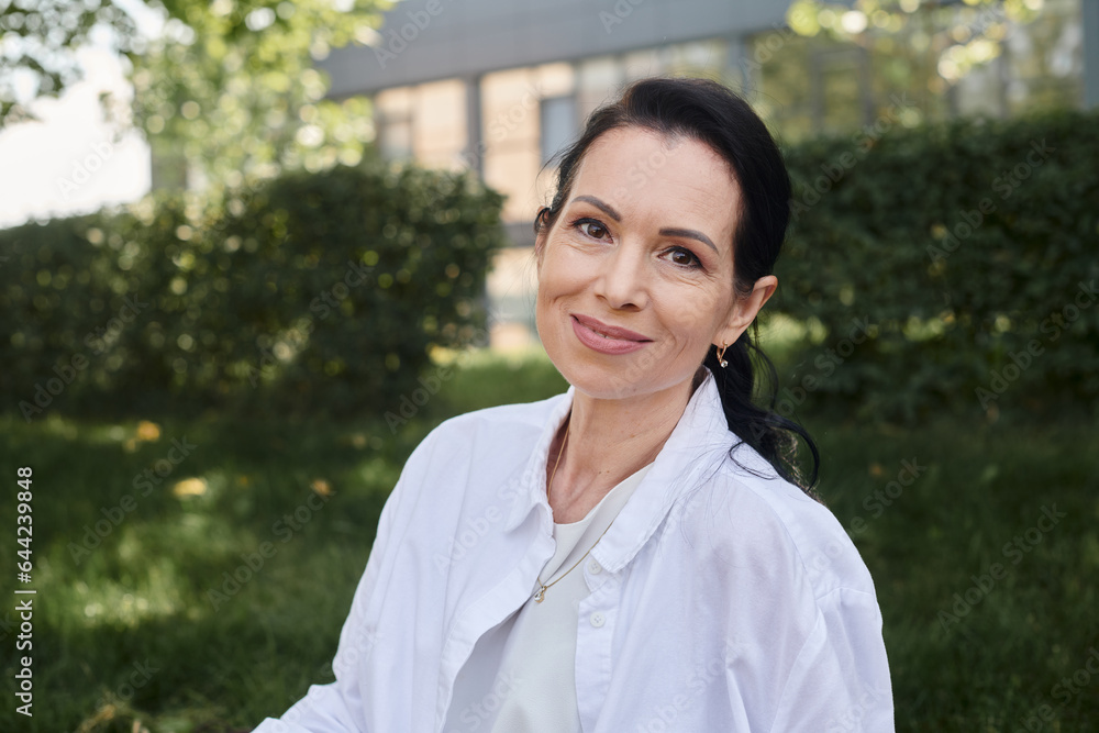 portrait of positive and stylish middle aged woman in white shirt smiling at camera in park portrait of positive and stylish middle aged woman in white shirt smiling at camera in park