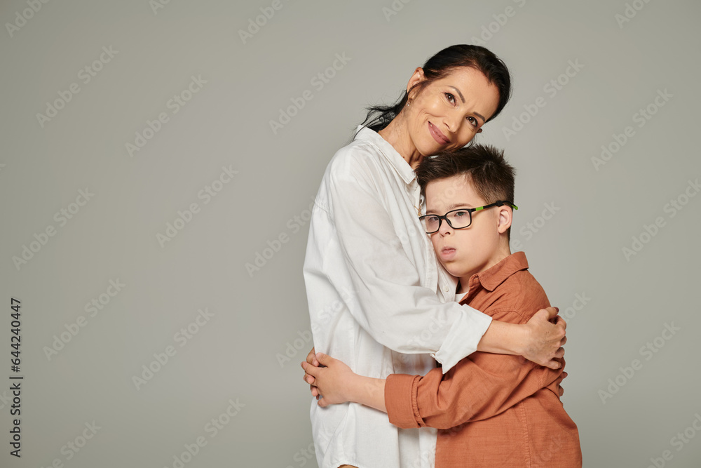 joyful middle aged woman hugging son with down syndrome and smiling at camera on grey, banner joyful middle aged woman hugging son with down syndrome and smiling at camera on grey, banner