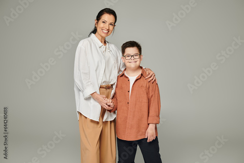 middle aged woman and son with down syndrome wearing stylish clothes and holding hands on grey