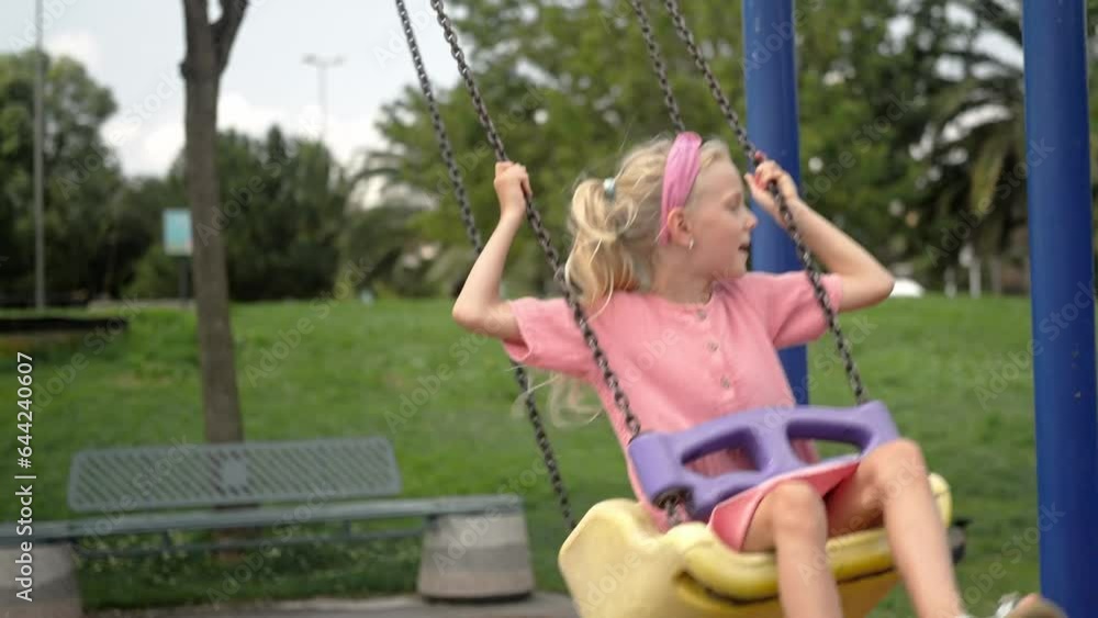 Blonde little girl rides swings in green city park cute child in pink dress enjoys swinging on children playground in garden kid plays attraction on gloomy day Blonde little girl rides swings in green city park cute child in pink dress enjoys swinging on children playground in garden kid plays attraction on gloomy day