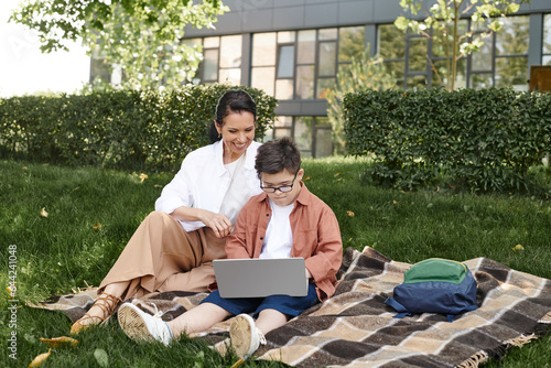 boy with down syndrome, in eyeglasses, sitting with laptop in park near happy mother, e-learning © LIGHTFIELD STUDIOS