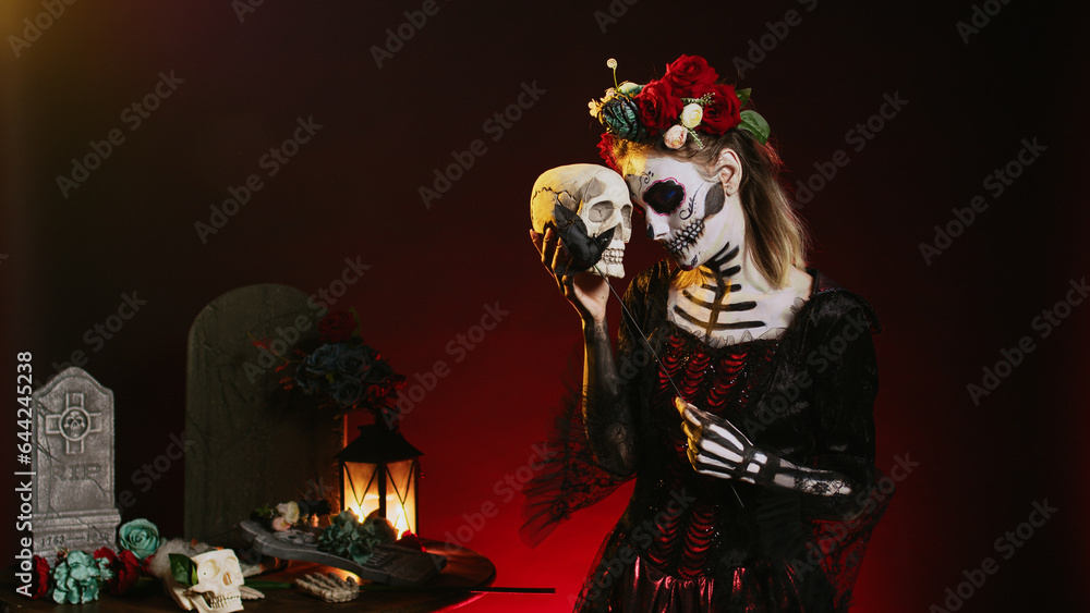 Spooky glamorous woman holding holy skull in studio, acting scary and horror to celebrate mexican halloween day. Flirty goddess wearing festival costume with body art, looking like lady of death.