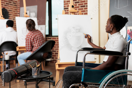 Disabled African American girl in wheelchair drawing on canvas during group art class, improving mental health through creativity. Person with disability learning to draw enjoying creative process