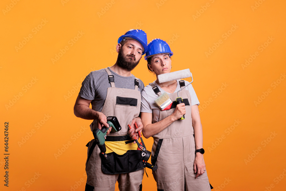 Exhausted team of contractors working with construction tools, falling asleep after renovating project. Man and woman builders holding power drill, painting brush or roller and pair of pliers.
