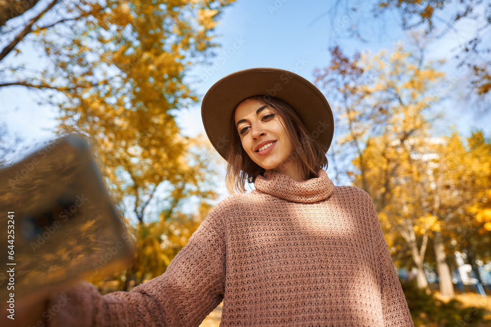 Woman in a chic hat and cozy knit sweater capturing perfect selfie moment on smartphone while strolling through sunny autumn park