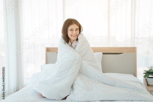 Photo of young happy woman in pajama stretching her arms and smiling while sitting on bed after sleep