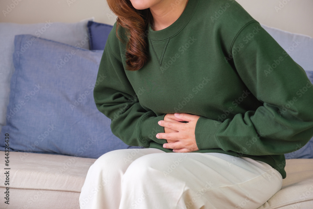 Closeup image of a sick woman suffering from stomachache, abdominal pain while sitting on sofa at home