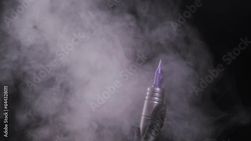 Female hand in black latex glove holds machine for permanent makeup and tattoos. In smoke. Concept of cosmetology procedures
