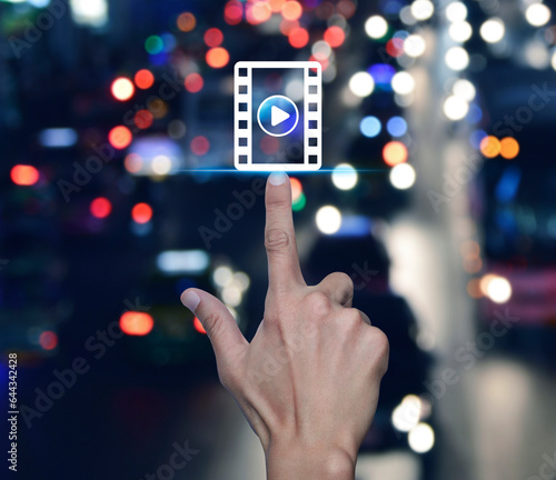 Hand pressing play button with movie flat icon over blur colourful night traffic jam road in city, Business cinema online concept