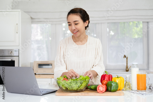 Happy young asian woman preparing food while following recipe on tablet laptop in the kitchen.