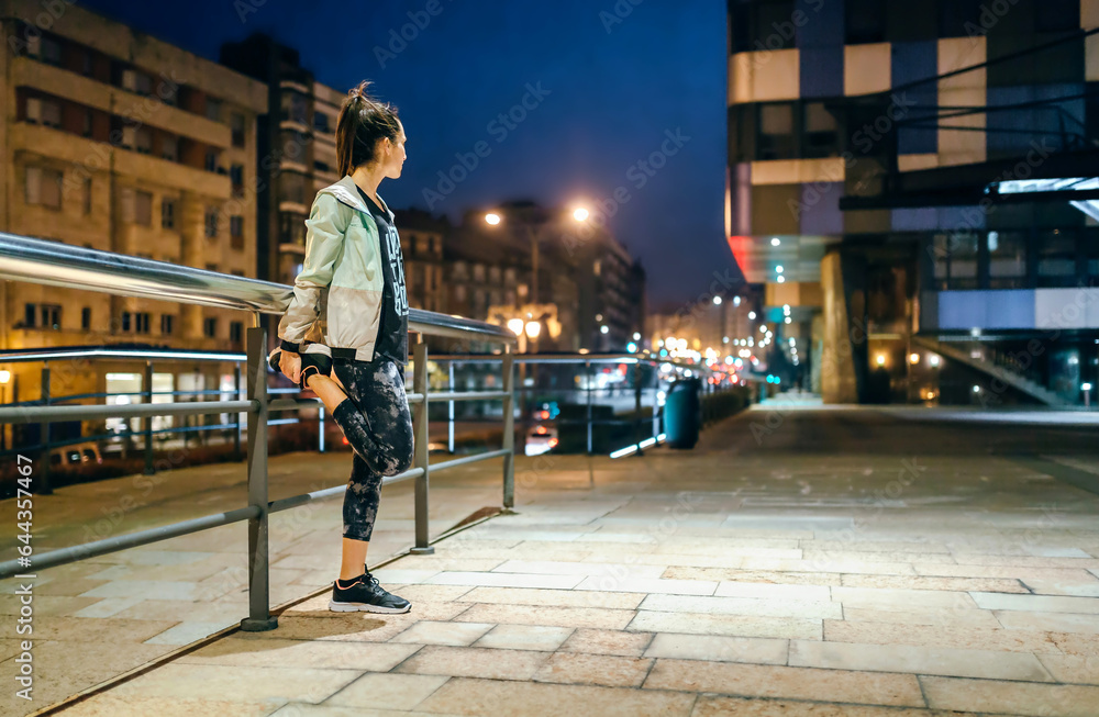 Unrecognizable young brunette woman runner with ponytail stretching her legs next to banister before training at night on empty town. Right copy space.