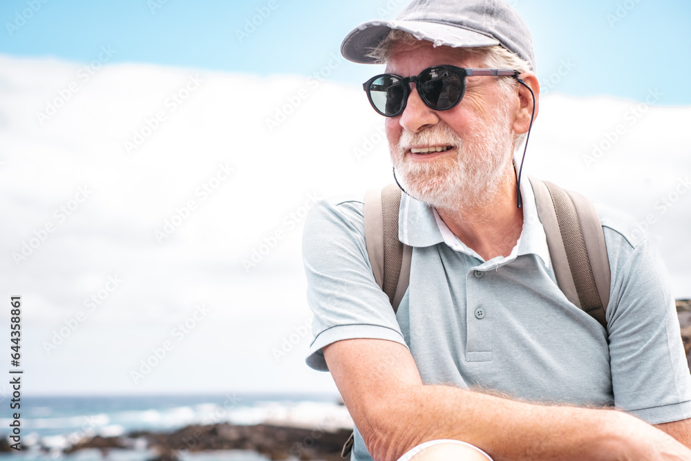 Portrait of smiling bearded senior man with cap and sunglasses sitting at sea carrying backpack enjoying summer vacation