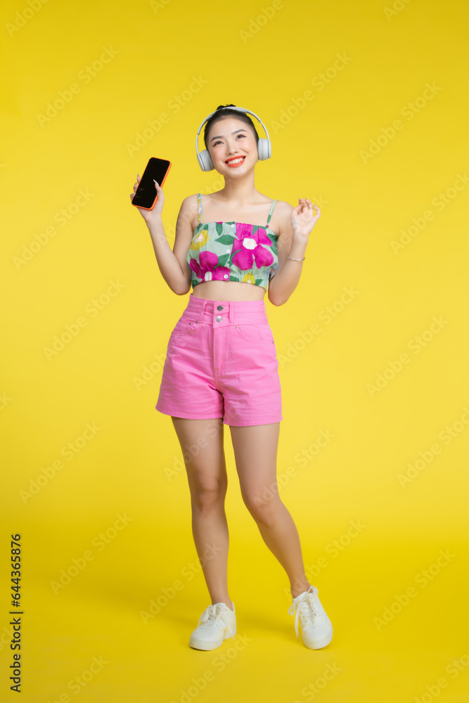 Asian pretty woman wearing headphones and holding on her cell phone over yellow background. Asian pretty woman wearing headphones and holding on her cell phone over yellow background.