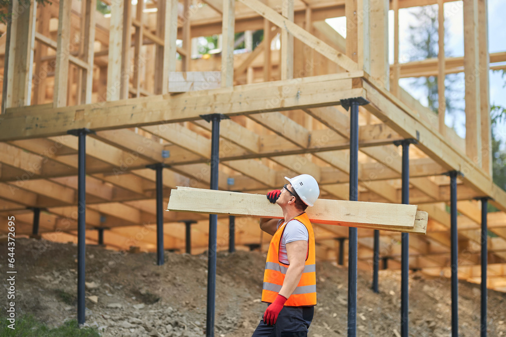 Side view of male builder wearing uniform and helmet, holding wooden boards, gilders, looking up. Man, worker building wooden house in forest in summer. Concept of building. Side view of male builder wearing uniform and helmet, holding wooden boards, gilders, looking up. Man, worker building wooden house in forest in summer. Concept of building.