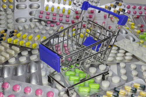 Empty decorative shopping cart stands on heap of multicolored pills, medicine shopping concept