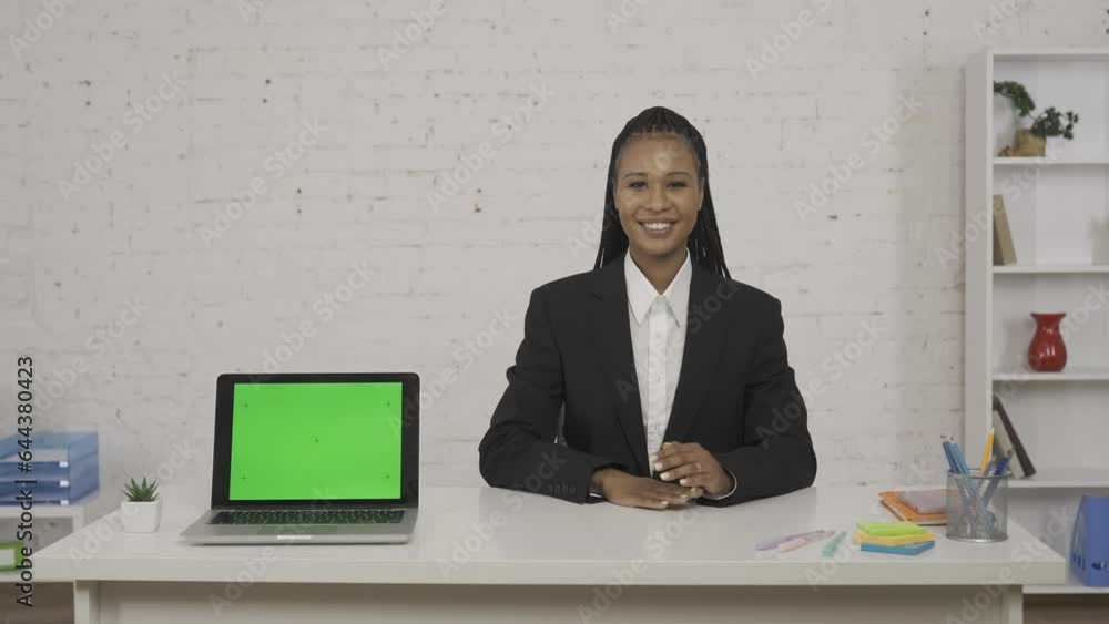 Modern businesswoman concept. Woman sitting at the desk smiling, pointing at the laptop next to her. Advertising area, workspace mockup. HDR BT2020 HLG Material. Modern businesswoman concept. Woman sitting at the desk smiling, pointing at the laptop next to her. Advertising area, workspace mockup. HDR BT2020 HLG Material.